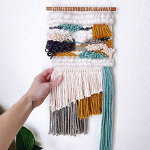 Load image into Gallery viewer, woven wall art hanging on a dowel with cream, teal, charcoal, and mustard fringe and details hand touching it to show size