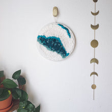 Load image into Gallery viewer, 12 " round handcrafted woven wall hanging with teal, aqua, blue surrounded by creamy white fibers hung from a brass hook.