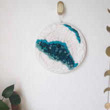 Load image into Gallery viewer, 12 " round handcrafted woven wall hanging with teal, aqua, blue surrounded by creamy white fibers hung from a brass hook.