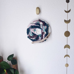 handcrafted 10" round woven wall hanging with lavender, mint, eggplant, teal and creamy white accented with driftwood and hung from a bronze hook.