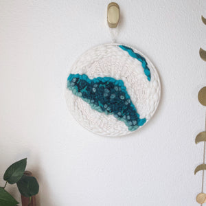 12 " round handcrafted woven wall hanging with teal, aqua, blue surrounded by creamy white fibers hung from a brass hook.