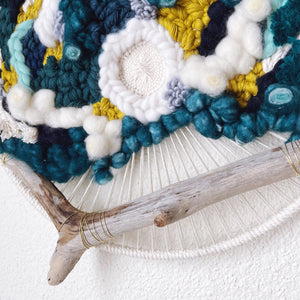 Close up of handcrafted 12" round wall hanging with teal, aqua, seafoam, navy, pistachio and creamy white natural fibers accented by driftwood